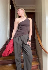 Brązowy top na jedno ramię zestawiony z luźnymi oliwkowymi spodniami. Modelka trzyma czerwoną skórzaną kurtkę i nosi na sobie stos bransoletek. W tle drewniane schody.