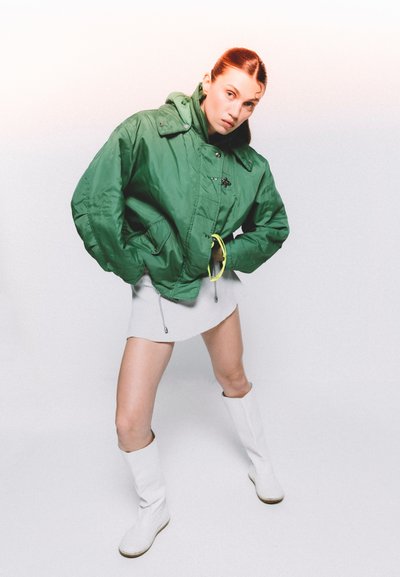 Chaqueta acolchada verde con cuello alto, presenta accesorios en negro, llevada sobre un vestido mini blanco. Modelo luciendo botas blancas hasta la rodilla.