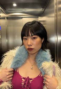 Personne aux cheveux noirs portant un haut violet et une veste blanche duveteuse avec col bleu, se tenant à l'intérieur d'un ascenseur en métal.