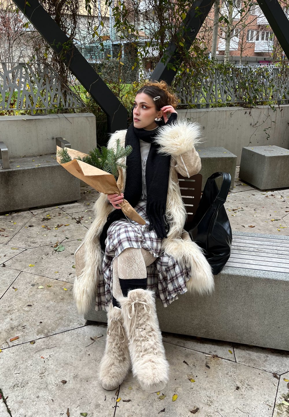 Donna seduta su una panchina di pietra che indossa un lungo cappotto in pelliccia sintetica color crema, sciarpa nera, gonna a scacchi e stivali beige pelosi, mentre tiene in mano della vegetazione avvolta.