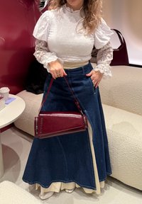 Top bianco con texture e maniche in pizzo, abbinato a una gonna di denim blu e una borsa in pelle rossa. Dettagli notevoli sui polsini delle maniche.