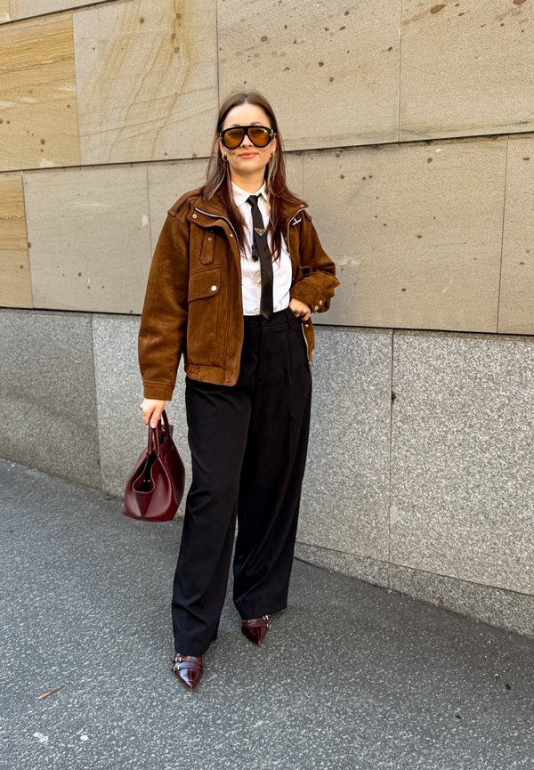 Donna che indossa una giacca di camoscio marrone, camicia bianca con cravatta nera, pantaloni neri, scarpe appuntite marroni, occhiali da sole, che tiene una borsa bordeaux sul marciapiede della città.