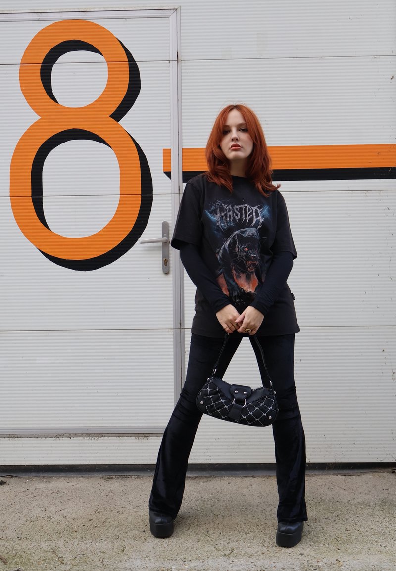 Jeune femme aux cheveux roux, portant un t-shirt graphique noir et un pantalon, se tient en tenant un sac à main noir devant une porte blanche avec un grand numéro orange 8.