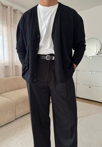 Mann, der einen schwarzen Cardigan, ein weißes T-Shirt, einen schwarzen Gürtel und schwarze Hosen trägt, steht mit den Händen in den Taschen in Innenräumen.