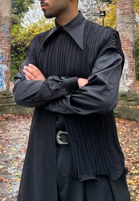 Homme portant une chemise noire plissée avec un grand col et les manches retroussées aux poignets, bras croisés, debout à l'extérieur près de piliers en briques.