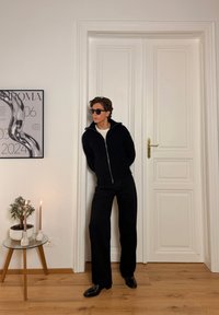 Pull en maille noir zippé, chemise blanche en dessous, pantalon large noir et chaussures noires. Décor minimaliste avec une plante et des bougies en arrière-plan.