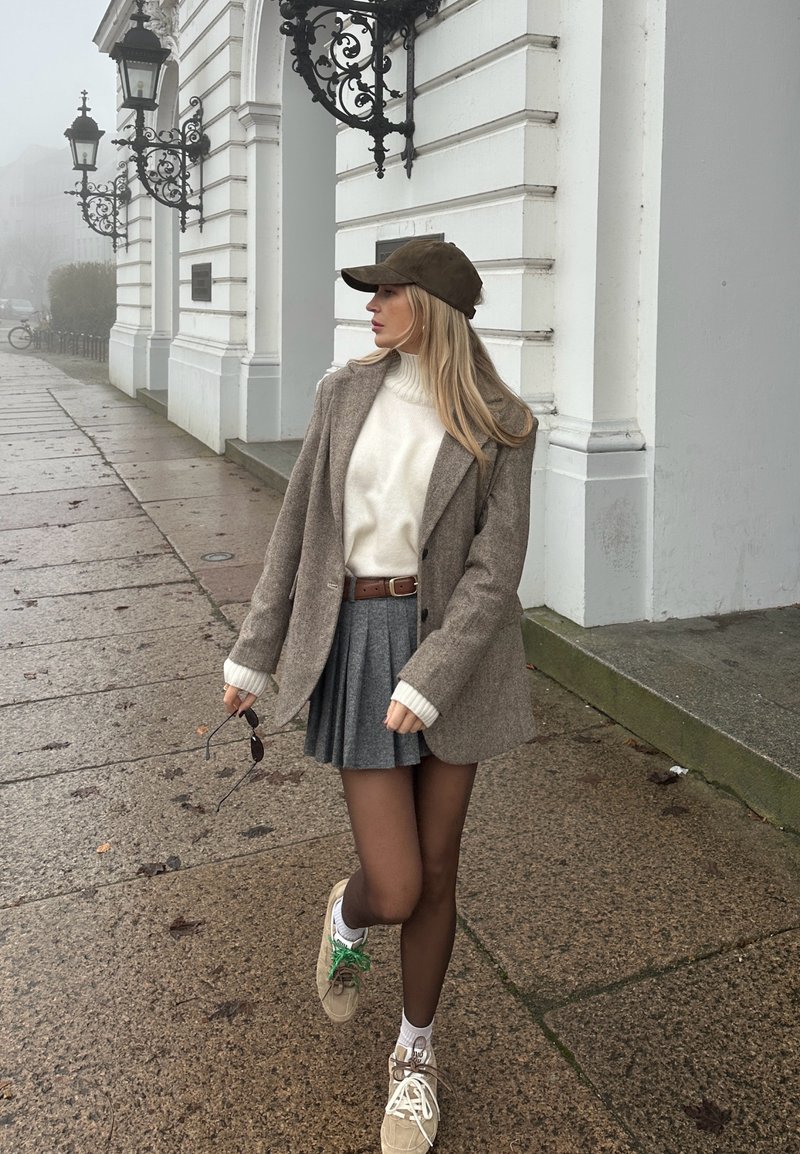Jeune femme portant une casquette marron, un blazer gris, un pull blanc, une jupe plissée grise et des baskets marchant sur un pavé de pierre près d'un bâtiment blanc.