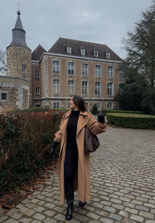 Frau in einem langen braunen Mantel, schwarzen Handschuhen und Stiefeln, die auf einem Kopfsteinpflasterweg in der Nähe eines historischen Steingebäudes mit Turm geht.