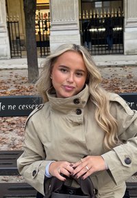 Beige trenssitakki, jossa tummat napit, päällä vaaleansininen paita. Pitkä, laineikas tukka, kädessä tumma käsilaukku ja sormissa kultaiset sormukset.