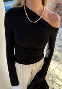 Top preto de manga longa com design assimétrico, textura franzida e decote off-the-shoulder, combinado com uma saia branca e colar de pérolas.