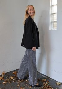 Mustast sobivast bleiserist, mis on istuv, koos hõbedaste litritega kaetud laiade säärega pükstega ja mustade madala kontsaga jalanõudega. Neutraalne taust.