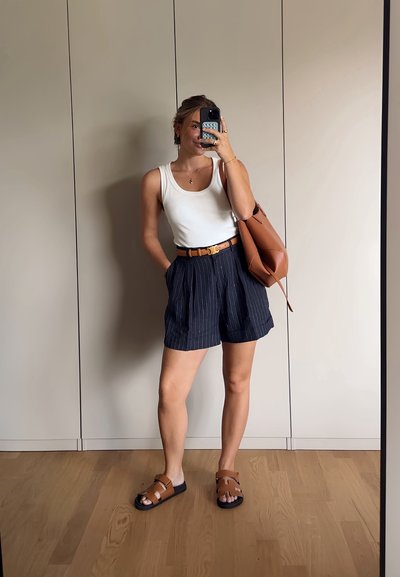 Camiseta sin mangas blanca, pantalones cortos de rayas marinas, cinturón marrón y sandalias marrones. Bolsa marrón con textura suave. Suelo de madera clara y fondo neutro.