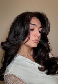 Giovane donna con lunghi capelli mossi e scuri, che indossa una maglietta bianca con maniche di pizzo e una delicata collana, che guarda in basso su uno sfondo semplice.