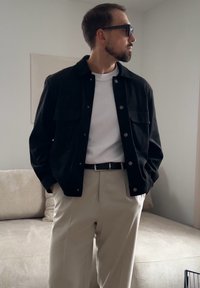 Άνδρας με μαύρο μπουφάν, λευκό πουκάμισο, μπεζ παντελόνι και γυαλιά ηλίου, στέκεται σε ένα σαλόνι με ουδέτερους τόνους με τα χέρια στις τσέπες.