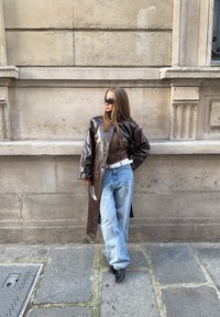 Femme portant un manteau en cuir brun foncé, une chemise à carreaux, un jean bleu clair, une ceinture blanche, des chaussures noires pointues et des lunettes de soleil, debout contre un mur en pierre.