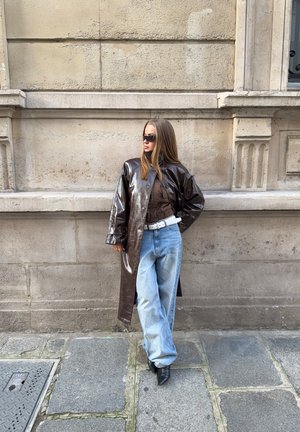 Mujer con abrigo de cuero marrón oscuro, camisa a cuadros, vaqueros azul claro, cinturón blanco, zapatos negros puntiagudos y gafas de sol, de pie junto a una pared de piedra.