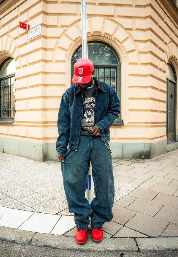 Persona con chaqueta vaquera y jeans de gran tamaño, gorra roja y zapatos, de pie en una esquina de la ciudad bajo una señal de calle cerca de un edificio beige.