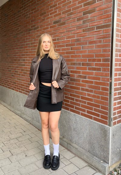 Chaqueta de cuero marrón sobre un top corto negro de canalé, falda negra, medias altas blancas y mocasines negros, con un fondo de pared de ladrillo.