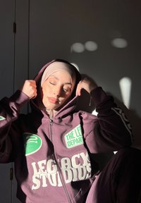 Sweatshirt bordeaux com grandes logótipos brancos e verdes, tecido texturizado, ajuste justo. A luz destaca as características do modelo e o discreto lenço bege na cabeça.