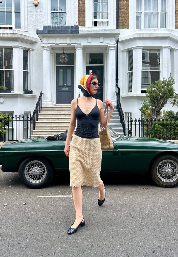 Donna con foulard in testa, occhiali da sole, top nero e gonna color crema cammina davanti a un'auto d'epoca verde e a una casa a schiera bianca.