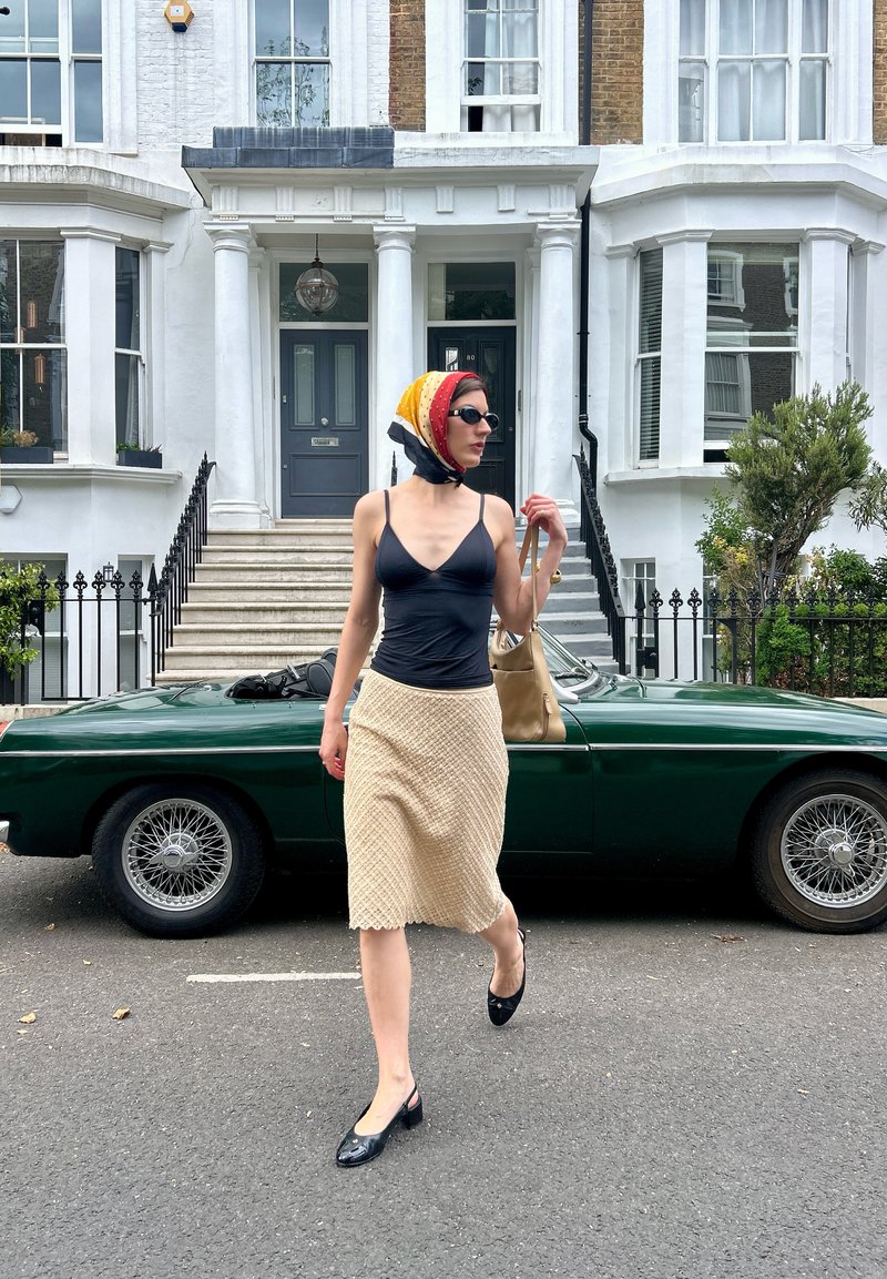 Femme portant un foulard, des lunettes de soleil, un haut noir et une jupe crème marche devant une voiture ancienne verte et une maison de ville blanche.