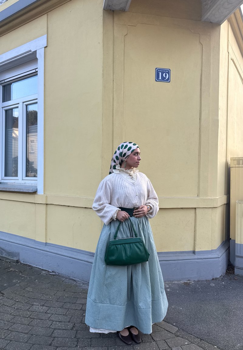 Femme vêtue d’un pull blanc, d’une jupe bleue, d’un foulard à pois sur la tête et d’un sac à main vert, debout à côté d’un bâtiment jaune avec une fenêtre et le numéro 19.
