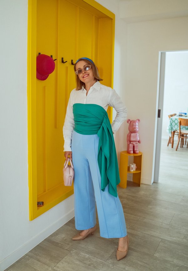 Femme portant des lunettes de soleil, une chemise blanche, un haut vert, un pantalon bleu et des talons beiges, debout près d'un panneau mural jaune avec un chapeau rouge suspendu.