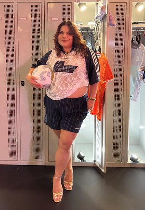 Camiseta de Adidas Originals con cordones y logo, pantalones cortos a rayas azul marino, sosteniendo un balón de fútbol. Fondo de vestuario con ropa deportiva y calzado.