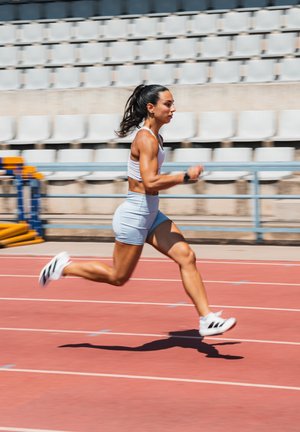 Atleta che corre su una pista rossa, indossando un reggiseno sportivo bianco e pantaloni corti azzurri chiari, con scarpe da ginnastica bianche con strisce nere. Sullo sfondo ci sono seggiolini vuoti nello stadio.