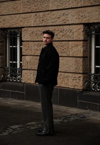 Svart knappet jakke over grå bukser, kombinert med svarte sko. Bakgrunnen har en teksturert steinvegg og et vindu med utsmykket metallarbeid.