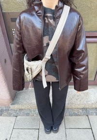Veste zippée en cuir synthétique marron, pull à motif camouflage gris, pantalon noir et sac bandoulière crème avec une lanière et un clip. Texture lisse, design moderne.