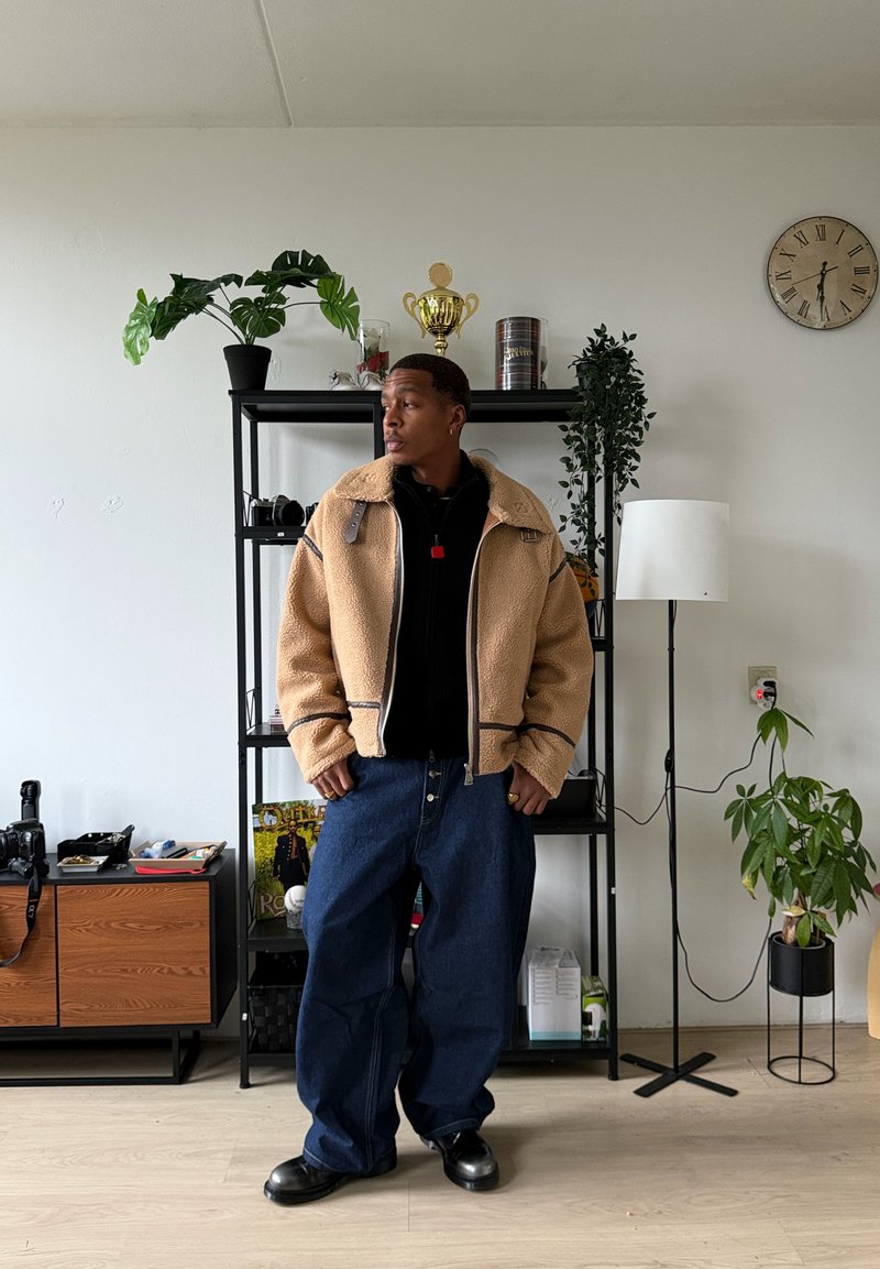 Veste en peau lainée beige avec fermeture éclair frontale, polo sombre en dessous, jean bleu foncé ample et chaussures noires. Cadre intérieur avec des plantes.