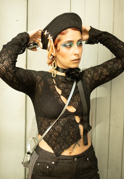 Top de encaje negro con cortes, combinado con pantalones de mezclilla negros de tiro alto. Los accesorios incluyen un sombrero negro y un bolso cruzado plateado.