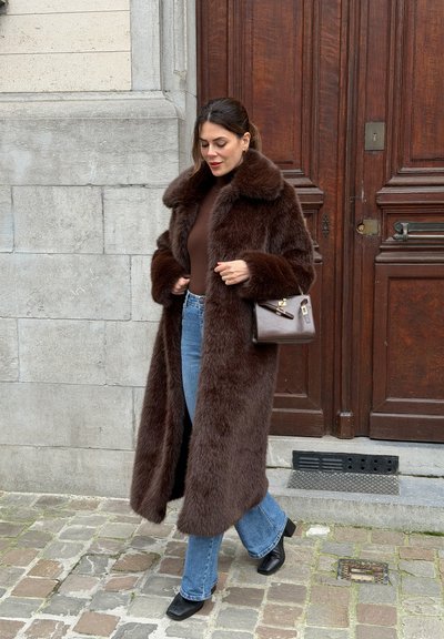 Abrigo marrón de piel sintética con un cuello ancho, combinado con una blusa marrón y jeans acampanados de mezclilla. Sostiene un pequeño bolso marrón; está de pie sobre un pavimento de adoquines.