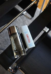 Klar glas parfume flaske med sølv låg og hvid TOCOBO solcreme flaske hvilende på sort stof stol.