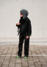 Černá kožená bunda s třásněmi, šedá kožešinová čepice, černé vzorované kalhoty a zelené boty s bílou podrážkou na kamenitém chodníku.