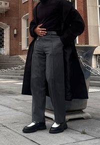 Manteau noir, col roulé noir ajusté, pantalon gris taille haute avec plis, et chaussures épaisses noires et blanches. Arrière-plan en milieu urbain.
