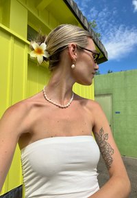 Top sem alças branco, colar de pérolas e brincos de pérolas. Cabelo em coque com uma flor branca. O fundo apresenta paredes amarelas e verdes brilhantes.