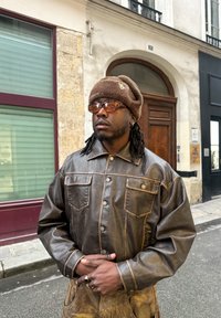 Καφέ δερμάτινο μπουφάν με ορατές ραφές και δύο τσέπες στο στήθος, συνδυασμένο με καφέ μπερέ και γυαλιά με φίλτρο, μπροστά από φόντο κτιρίου.