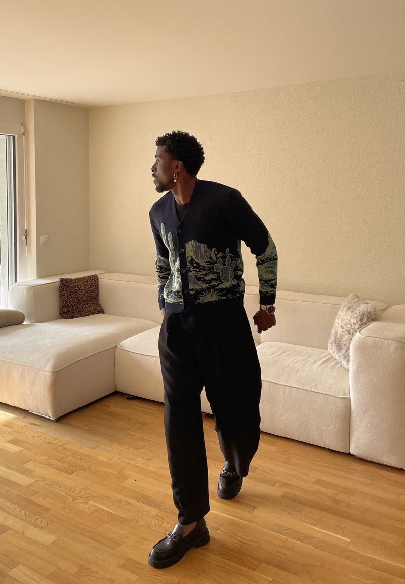 Homme en pantalon noir et cardigan à motif cactus marchant dans un salon lumineux avec un canapé sectionnel beige et un sol en bois.