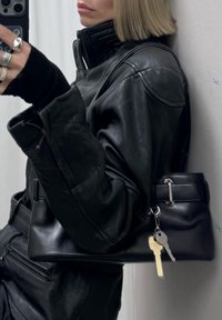 Femme en veste en cuir noir tenant un sac à main noir avec des clés attachées, prenant un selfie dans un miroir avec un smartphone.