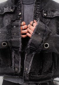 Black denim jacket with frayed edges, silver snap buttons, and grommets. Layered over a grey ribbed sweater. Hands adorned with various rings.