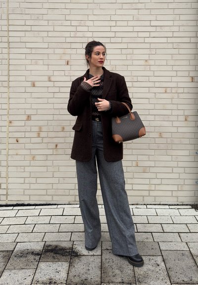 Mujer con un abrigo marrón oscuro, pantalones anchos grises, sosteniendo un bolso marrón, de pie sobre un suelo de baldosas frente a una pared de ladrillos claros.