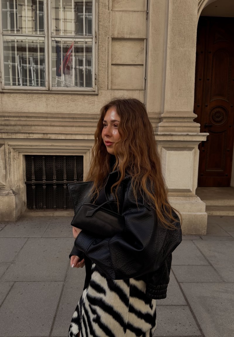 Femme aux longs cheveux ondulés marchant sur un trottoir en ville, portant une veste en cuir noire, une jupe à motifs zébrés, et tenant un sac à main noir près d'un bâtiment en pierre.
