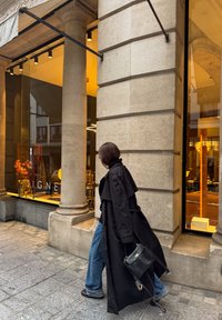 Čierny trenčkot, voľné modré džínsy a čierna kabelka. Kamenné stĺpy a osvetlená výkladná skriňa s viditeľným nábytkom a dekoráciami.