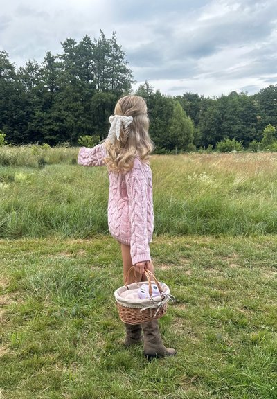 Suéter de punto trenzado color rosa claro, pantalones cortos casuales, botas marrones. Sosteniendo una cesta de mimbre con forro crema. Campo verde exuberante de fondo.