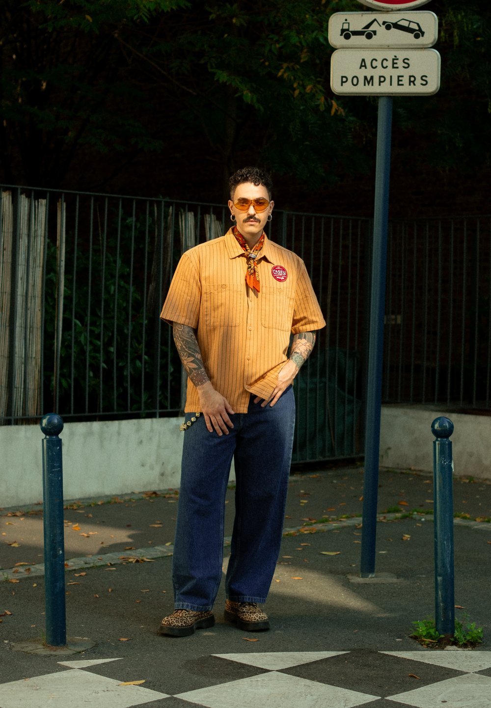 Uomo con tatuaggi che indossa una camicia arancione, jeans blu, scarpe leopardate e occhiali da sole arancioni mentre si trova vicino a un cartello di accesso al fuoco sulla strada.