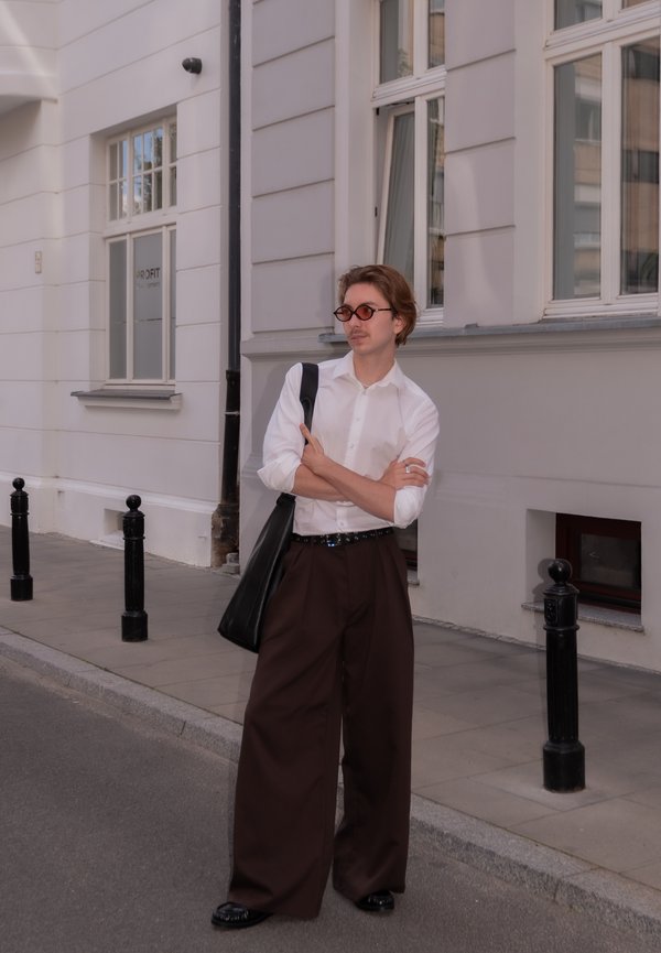 Hombre con gafas de sol redondas, camisa blanca y pantalones anchos marrones, de pie en una calle de la ciudad con los brazos cruzados y un bolso negro al hombro.