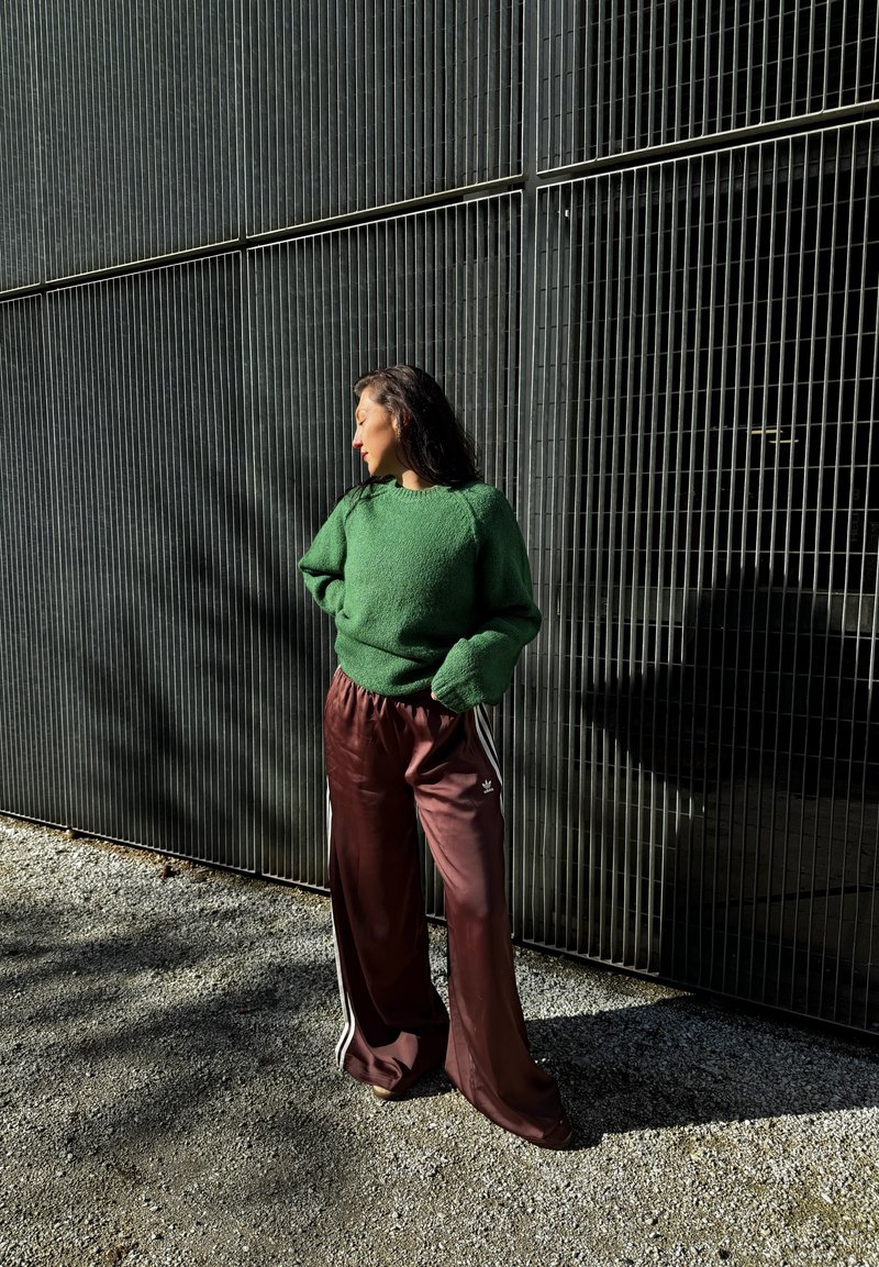 Suéter de textura verde combinado con pantalones anchos de satén burdeos con rayas blancas, de pie frente a un oscuro fondo con patrón de rejilla.