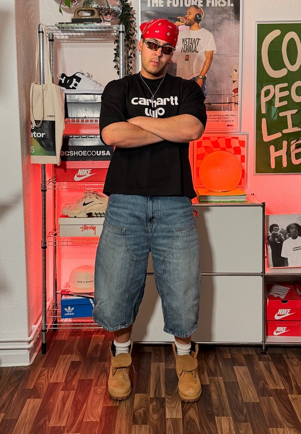 Mand med korslagte arme iført solbriller, rød bandana, sort Carhartt-shirt og denimshorts står i et rum med skohylder og plakater.